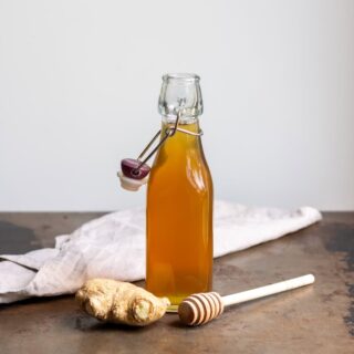 A bottle of honey ginger syrup on a table, next to a piece of fresh ginger and a wooden honey spoon.