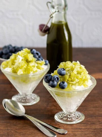 Side view of two glasses of kakigori Japanese shaved ice, topped with matcha syrup and blueberries.