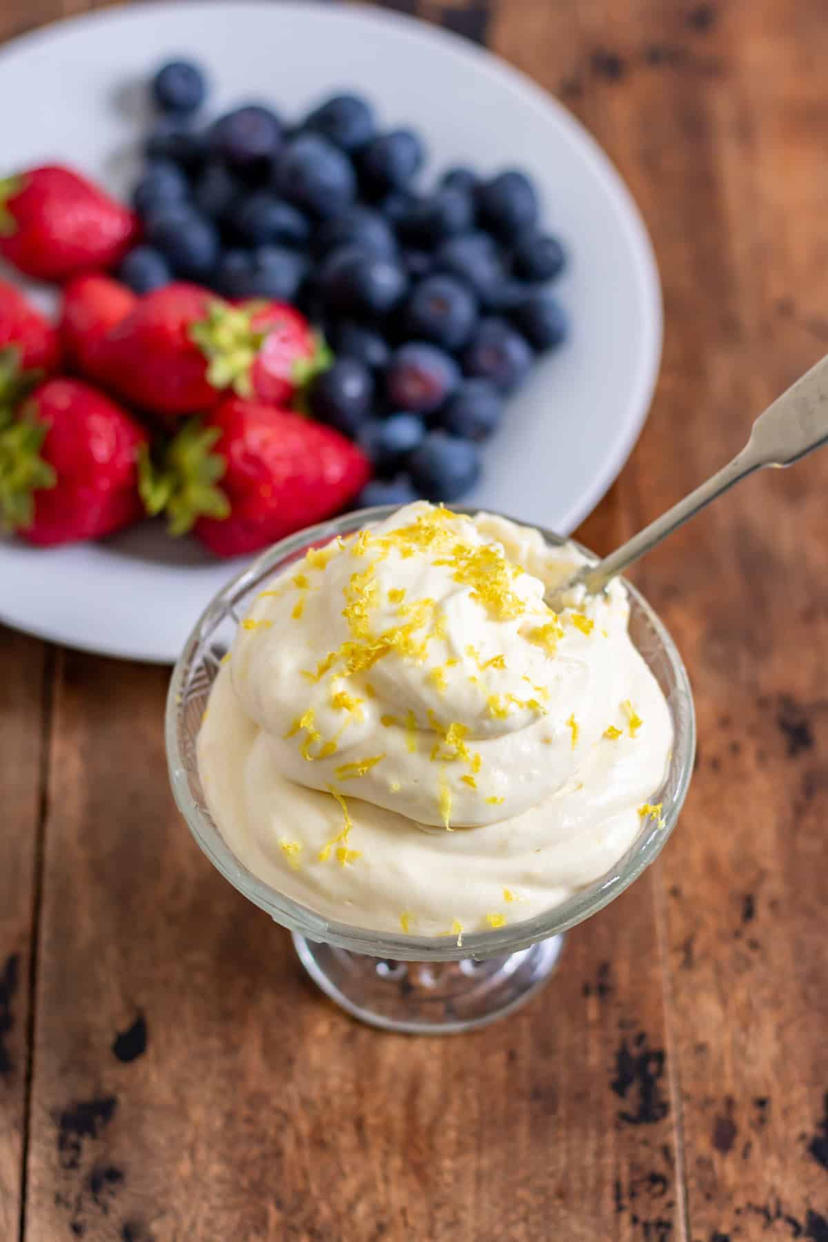 A glass dish piled with lemon whipped cream, topped with extra lemon zest, in front of a dish of berries.