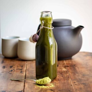 Wooden table with a spoon of matcha powder, bottle of matcha syrup and in the background a teapot and Japanese cups.