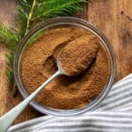 Spoon in a glass bowl of Lebkuchengewürz on a wooden table next to a tea towel and a sprig of pine.