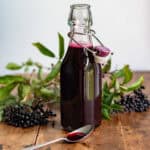 Bottle of elderberry syrup, in front of bunches of elderberries, plus a teaspoon of the syrup.