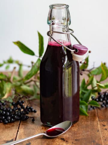 Bottle of elderberry syrup, in front of bunches of elderberries, plus a teaspoon of the syrup.