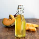 A bottle of melon compote on a wooden table, in front of slices of cantaloupe.