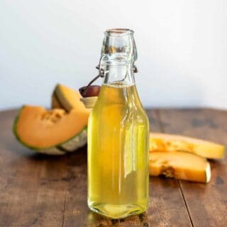 A bottle of melon compote on a wooden table, in front of slices of cantaloupe.