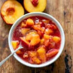 Wooden table with a small dish of nectarine compote with a spoon in it, and a cut nectarine next to it.