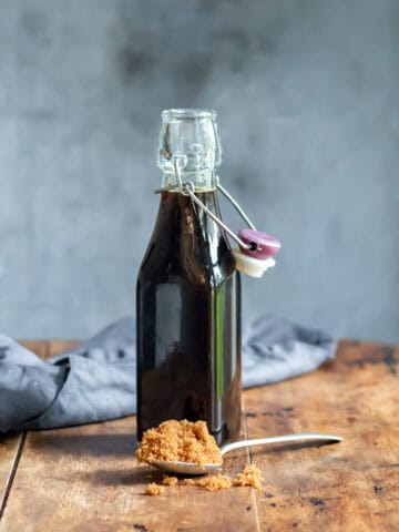 Wooden table with a glass bottle of brown sugar syrup with a teaspoon of brown sugar in front of it.