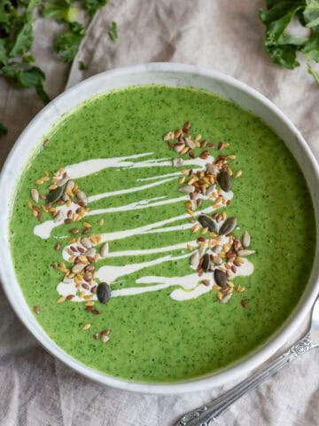 A table with a bowl of kale soup, drizzled with cream and sprinkled with seeds, on a napkin, surrounded by kale.