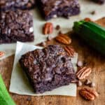 A square of vegan zucchini brownies on a piece of baking paper on a wooden table, next to courgettes and pecans.