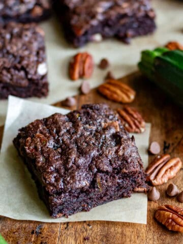 A square of vegan zucchini brownies on a piece of baking paper on a wooden table, next to courgettes and pecans.