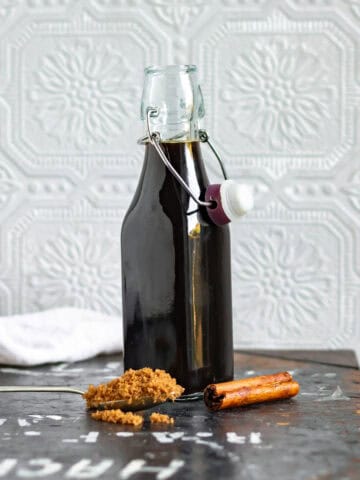 A glass bottle of cinnamon brown sugar syrup with a teaspoon of sugar and a cinnamon stick in front of it.