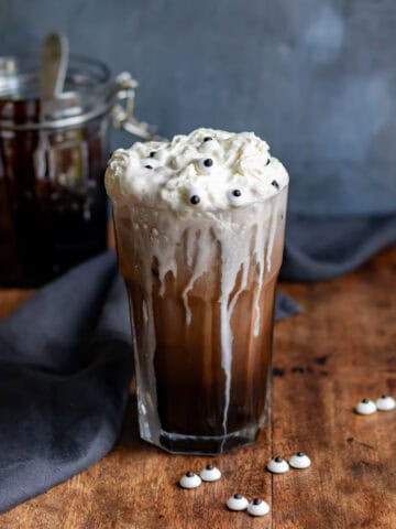 A glass of Halloween chocolate cream soda, topped with whipped cream and candy eyes, with cream drizzling down the outside of the glass.