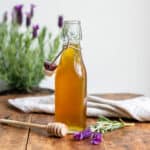 Glass bottle of lavender honey syrup on a wooden table, next to a wooden honey spoon and sprigs of lavender.
