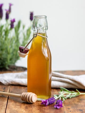 Glass bottle of lavender honey syrup on a wooden table, next to a wooden honey spoon and sprigs of lavender.