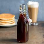 A glass bottle of pumpkin spice syrup on a table in front of a stack of pancakes and a latte.