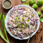 A wooden table with a dish of brussels sprout slaw, with a dish of pepper, two spring onions and some sprouts around it.