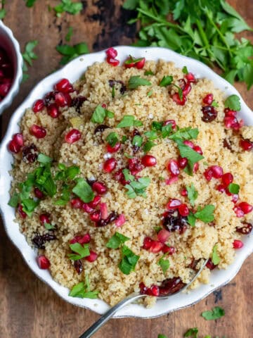 A wooden table with a bowl of Christmas couscous, next to a dish of pomegranate seeds and parsley.