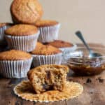 A wooden table with a pile of muffins, bowl of mincemeat, and a mincemeat muffin with a bite out in front.