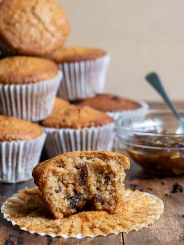 A wooden table with a pile of muffins, bowl of mincemeat, and a mincemeat muffin with a bite out in front.