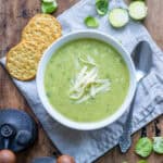 A wooden table with a bowl of Brussels sprout soup on a napkin surrounded by crackers and brussels sprouts.