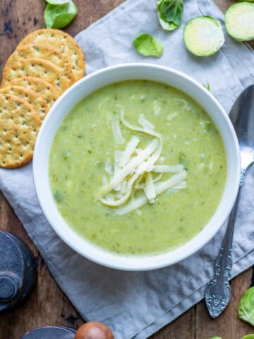 A wooden table with a bowl of Brussels sprout soup on a napkin surrounded by crackers and brussels sprouts.