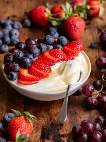 Side view of a bowl of whipped cottage cheese with a spoon in it, topped with sliced strawberries, blueberries and grapes, and a drizzle of honey.