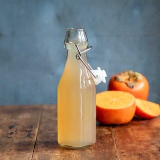 A bottle of persimmon syrup on a table in front of cut persimmons.