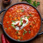 A wooden table with a serving dish of tomato lentil soup, topped with yogurt and harissa paste.