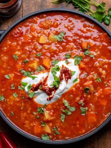 A wooden table with a serving dish of tomato lentil soup, topped with yogurt and harissa paste.