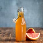 Wooden table with a glass bottle of grapefruit syrup next to slices of pink grapefruit.