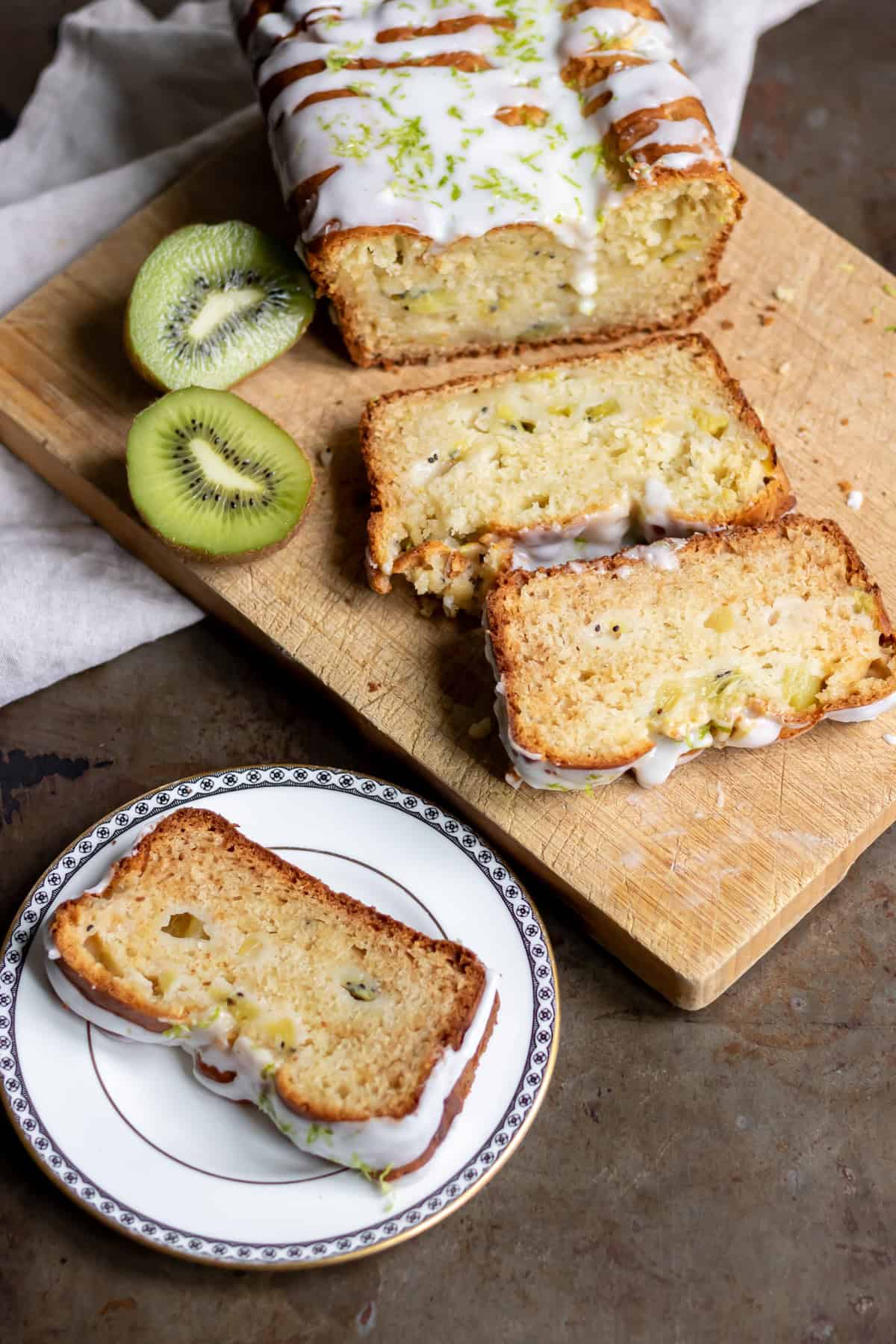 Looking down at a table with a slice of cake, and the kiwi loaf cake being cut on a wooden board.