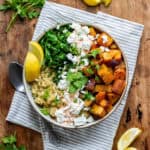 A wooden table with a bowl of roasted vegetable quinoa salad, with parsley and sliced lemons scattered around.