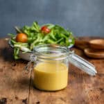 Wooden table with a jar of maple dijon dressing in front of a bowl of salad.