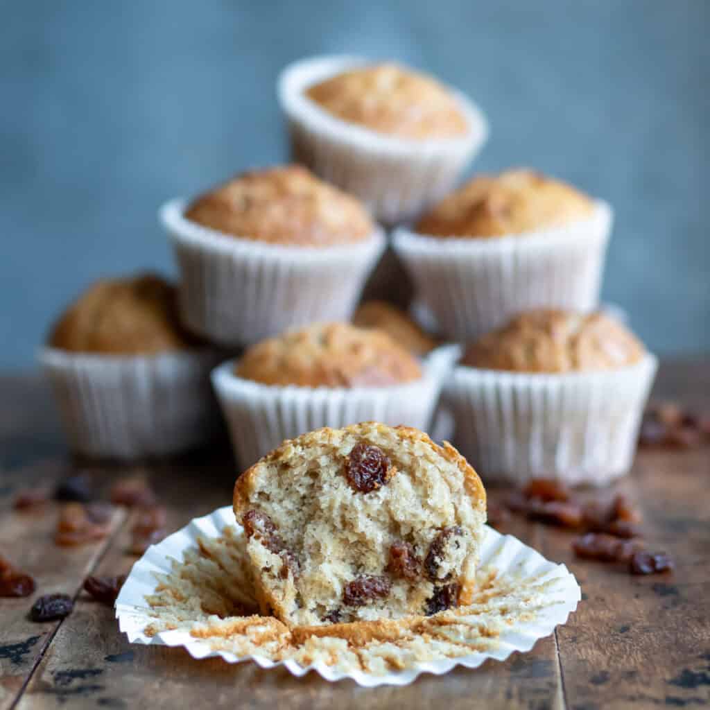 Side view of a table with a raisin muffin with a bite out in front of a pile of muffins.