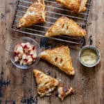 A wooden table with a rack of cooling nectarine turnovers, a few turnovers on the table, a dish of nectarines and a dish of coarse sugar.