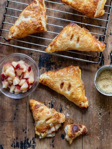 A wooden table with a rack of cooling nectarine turnovers, a few turnovers on the table, a dish of nectarines and a dish of coarse sugar.