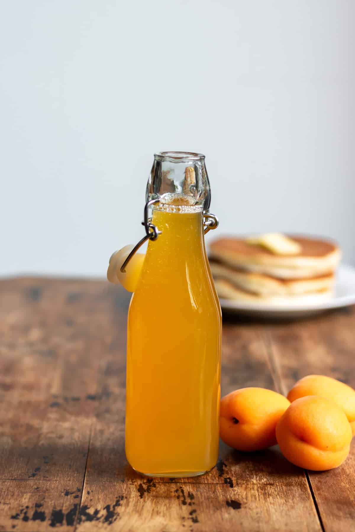 Wooden table with a stack of pancakes, apricots and bottle of apricot simple syrup.