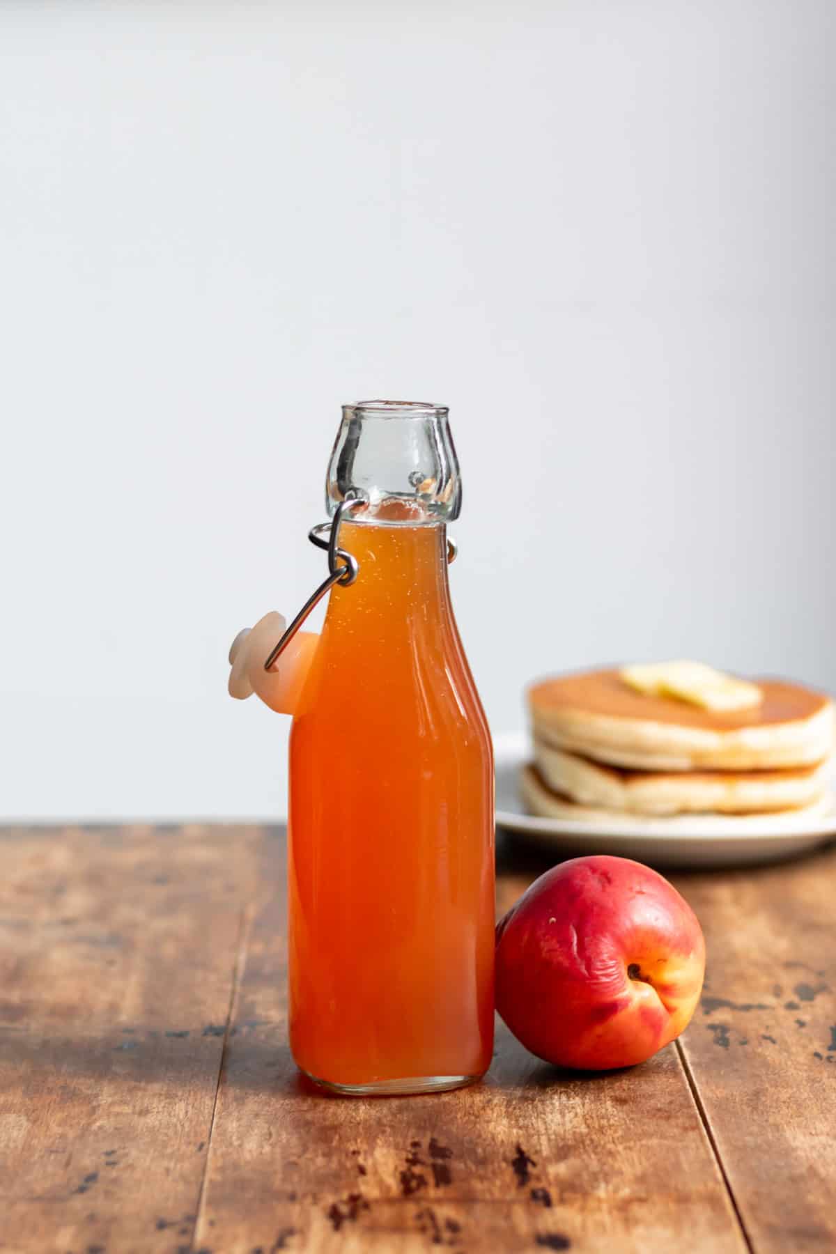 A glass bottle of nectarine simple syrup on a table in front of pancakes.