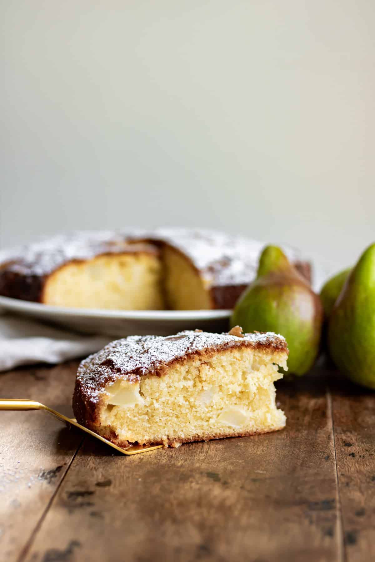 A table with a pear cake and a slice of it in the front next to pears.