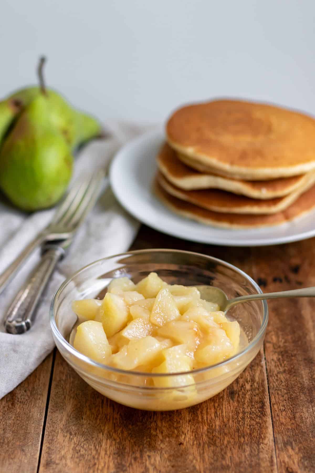 Side view of a glass dish of pear compote in front of a stack of pancakes.