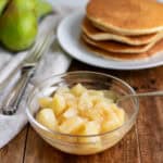 Side view of a glass dish of pear compote in front of a stack of pancakes.