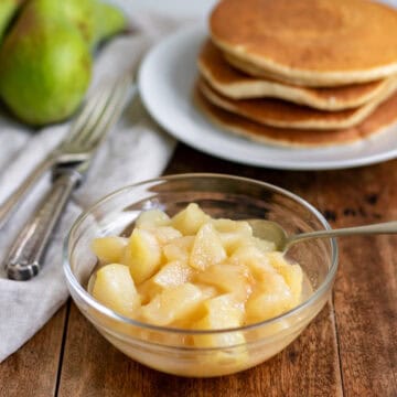 Side view of a glass dish of pear compote in front of a stack of pancakes.