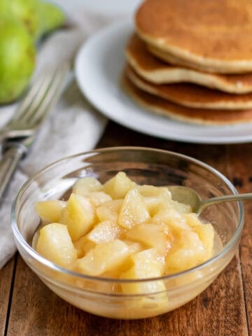 Side view of a glass dish of pear compote in front of a stack of pancakes.