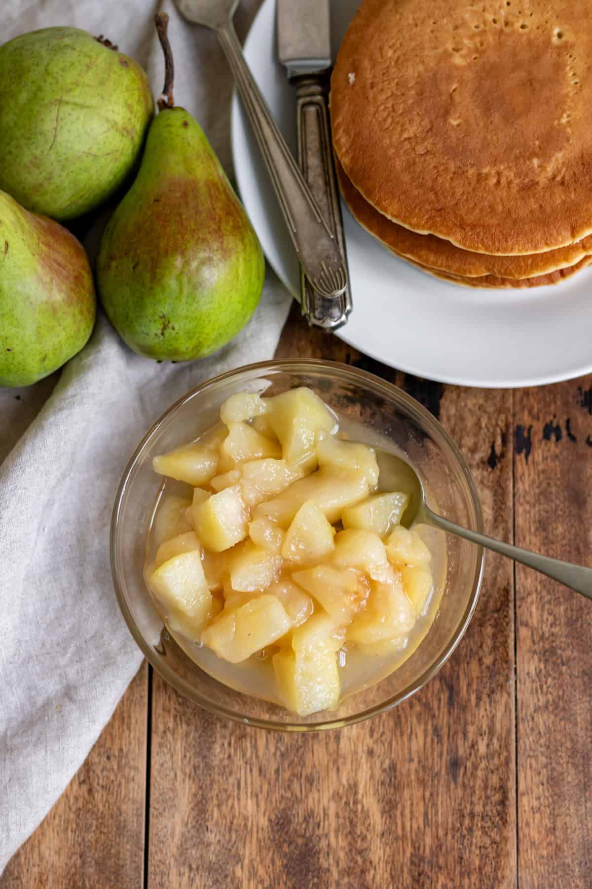 Looking down at a wooden table with a dish of pear compote, napkin, pears and a stack of pancakes.