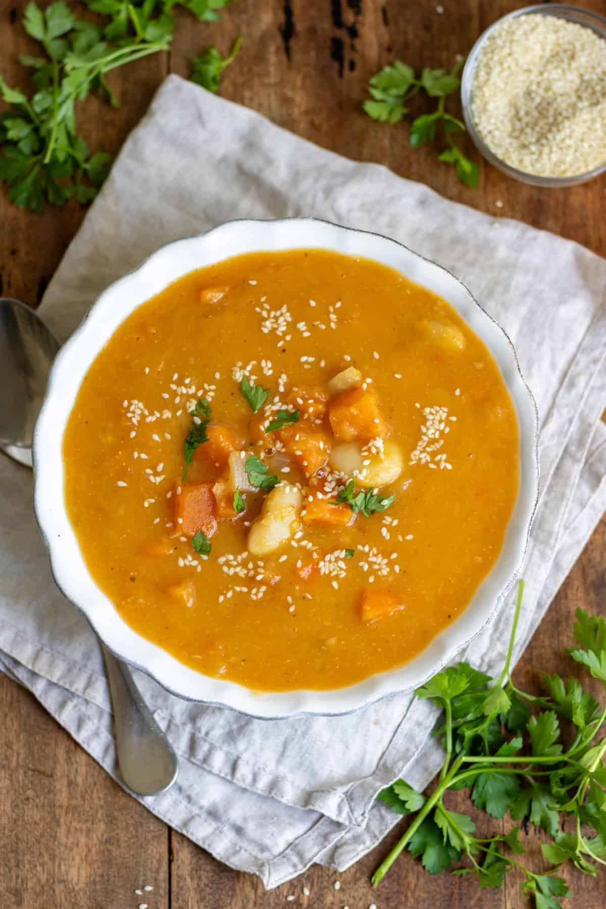 A table with a bowl of miso sweet potato soup on a napkin, next to fresh herbs and a dish of sesame seeds for topping.
