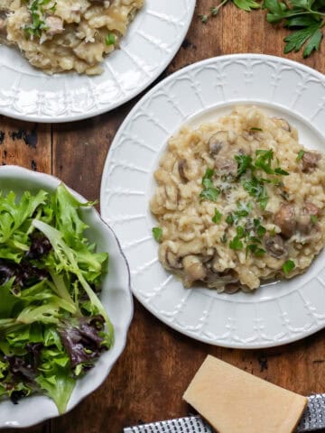 Wooden table with two plates of slow cooker mushroom risotto and a dish of salad.