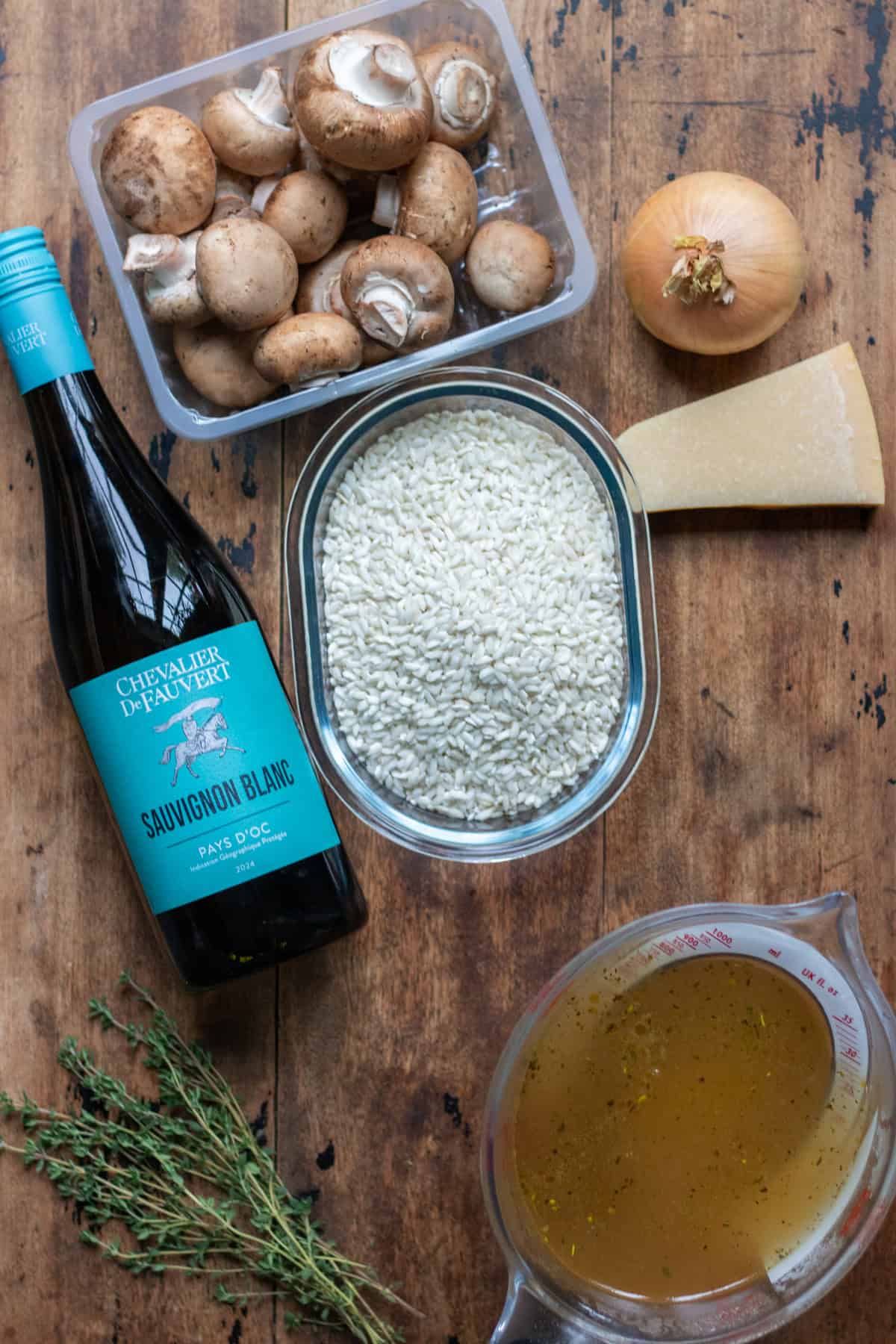 Mushrooms, onion, stock, thyme, parmesan, arborio rice and white wine, on a wooden table.
