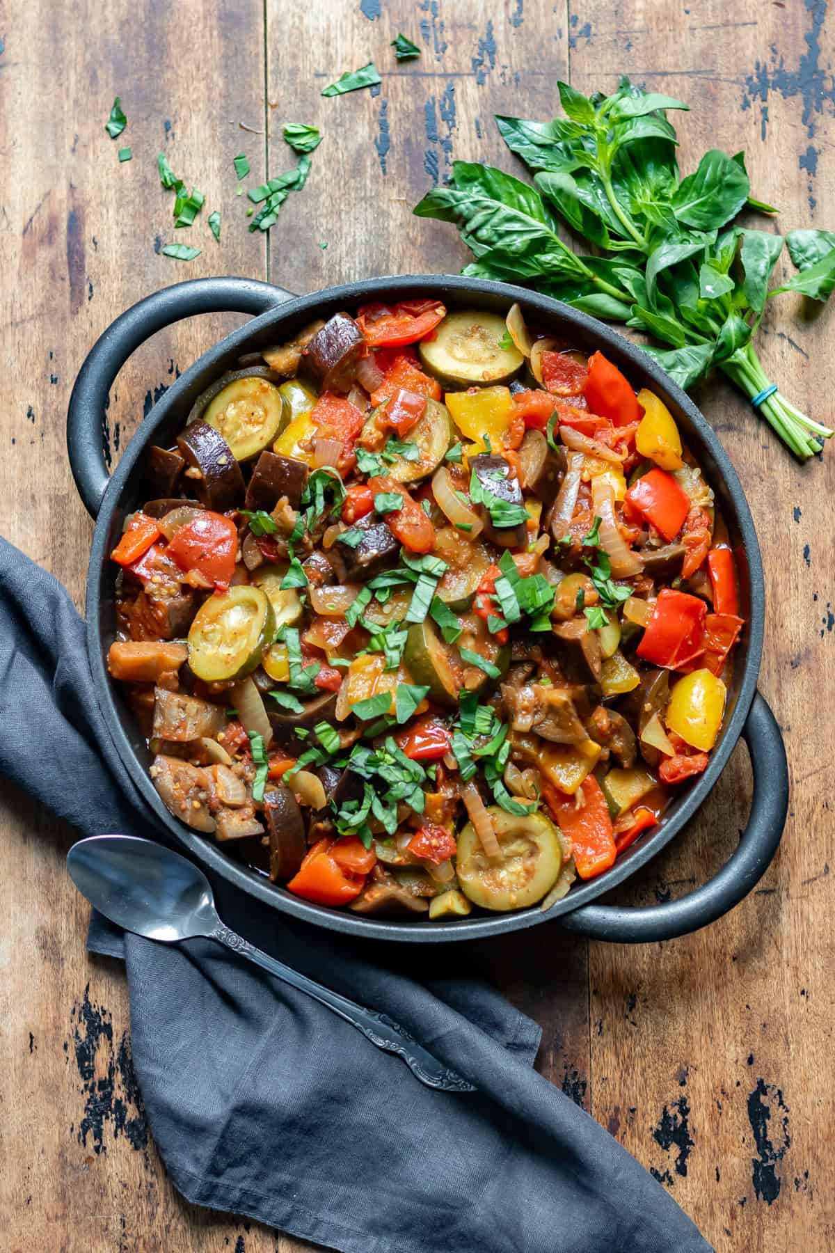 Wooden table with a cast iron serving dish of slow cooker ratatouille, next to a bunch of basil and a napkin.