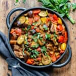 Wooden table with a cast iron serving dish of slow cooker ratatouille, next to a bunch of basil and a napkin.