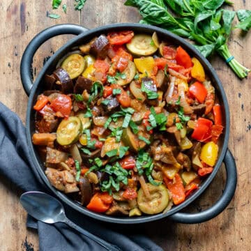 Wooden table with a cast iron serving dish of slow cooker ratatouille, next to a bunch of basil and a napkin.
